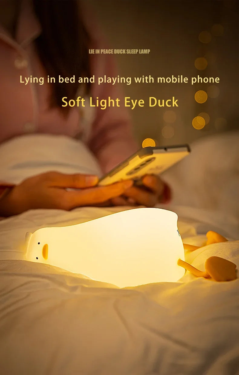 A soft light eye duck night light illuminates a hand holding a mobile phone in a bedroom setting.