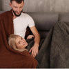 A man and a woman relaxing together, sharing a cozy waterproof throw blanket on a sofa.