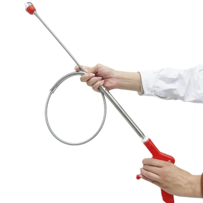 A hand is operating a drain unclogging tool with an ergonomic handle, showing the long metal probe and coiled end.