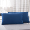 A set of two deep blue microfiber pillowcases resting on a gray bed, with a white and gray checkered curtain in the backgroun
