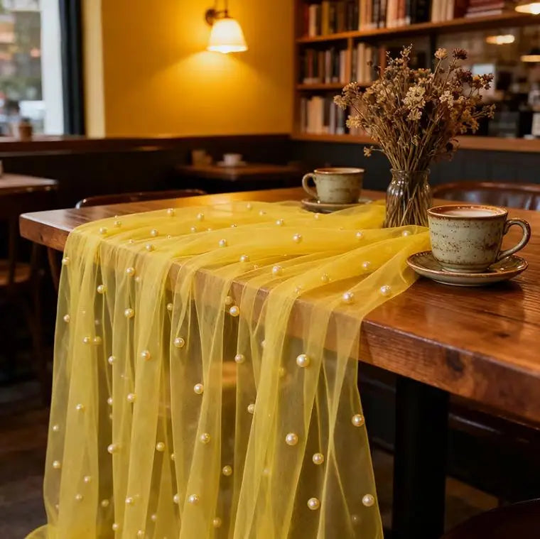 A sheer yellow pearl beaded tulle table runner draped over a wooden table in an indoor setting.