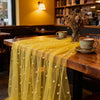 A sheer yellow pearl beaded tulle table runner draped over a wooden table in an indoor setting.