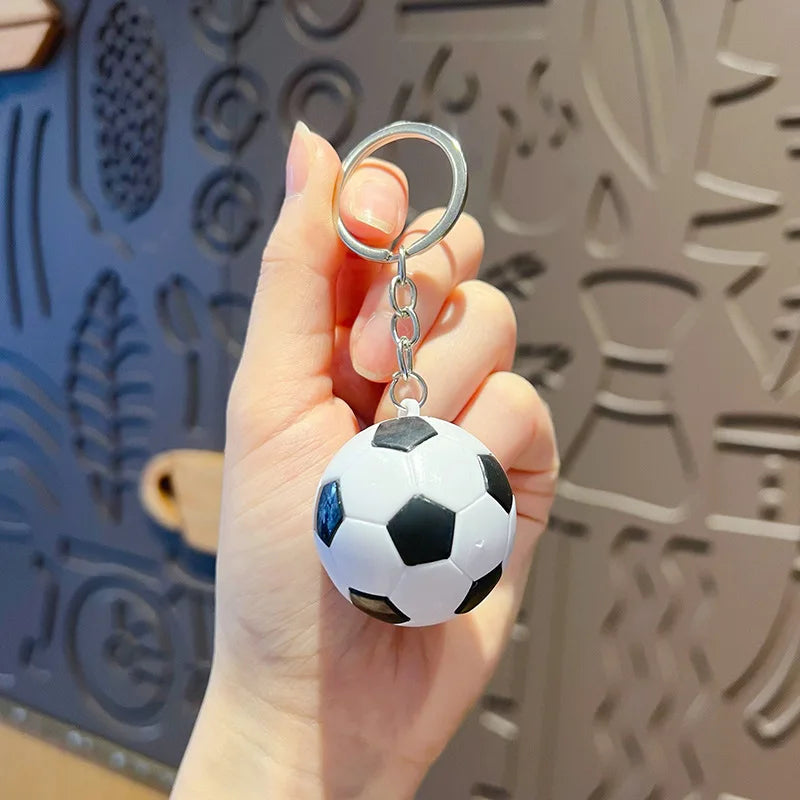 Una mano sostiene un llavero de balón de fútbol blanco y negro metálico contra un fondo estampado.