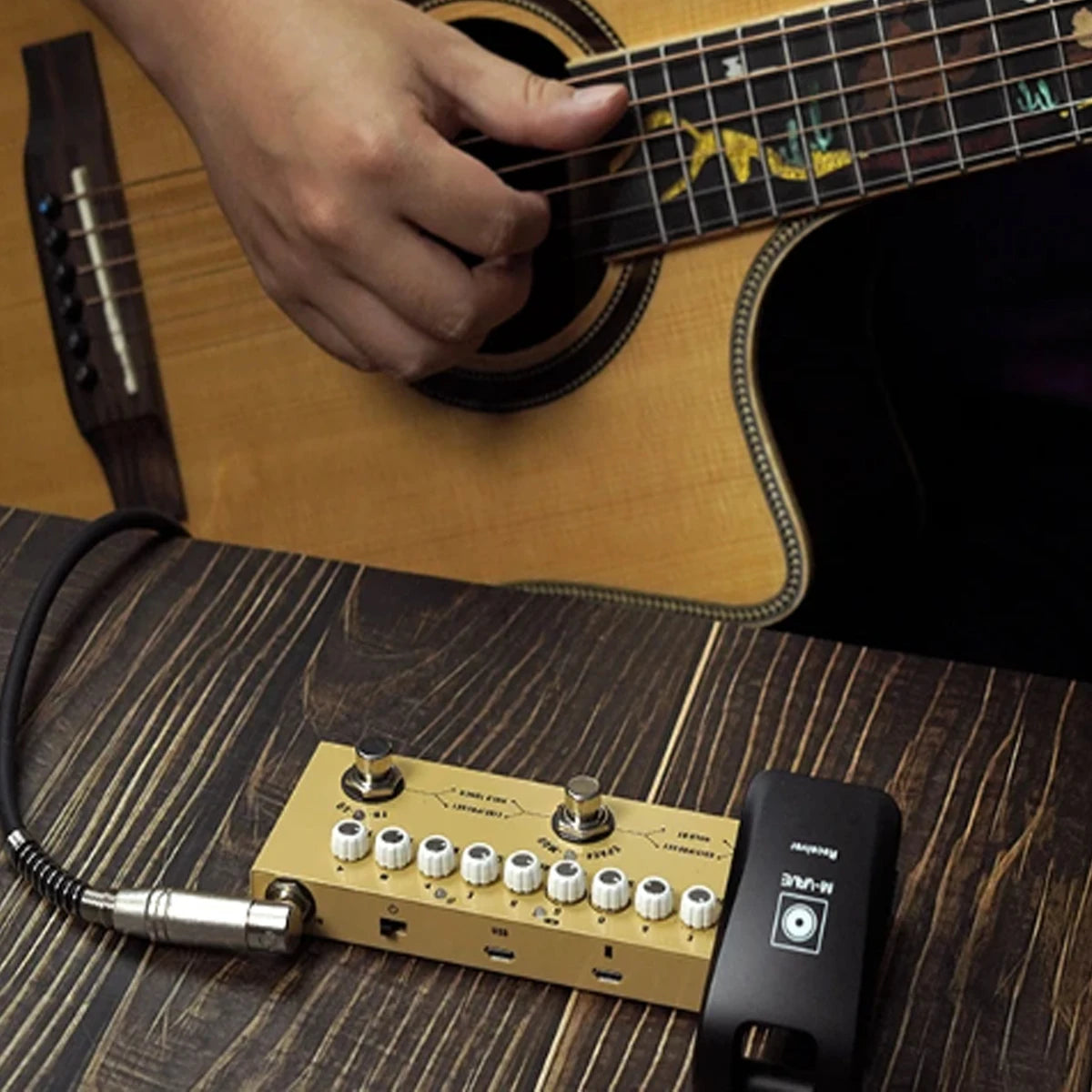 A close-up shot of a beige guitar pedal with multiple knobs and switches resting on a wooden surface next to an acoustic guit