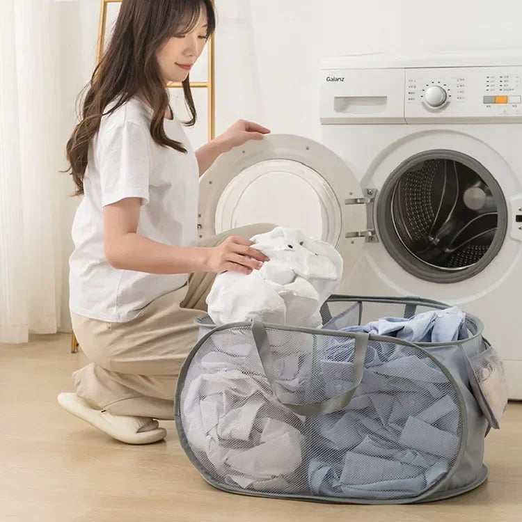 A woman is folding laundry in a gray mesh basket with handles next to a white washing machine.