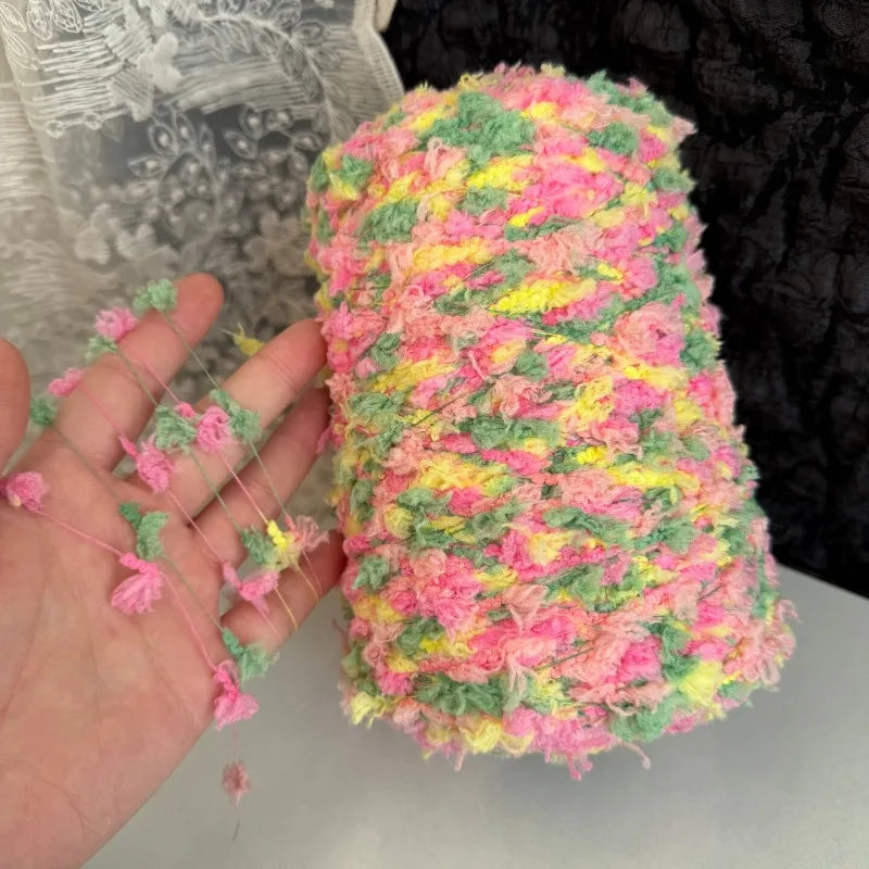 A hand is holding a skein of hand-dyed variegated cotton blend yarn featuring pink, yellow, and green fibers.