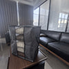 A dark gray foldable storage bag is displayed on a wooden surface next to a black leather sofa.