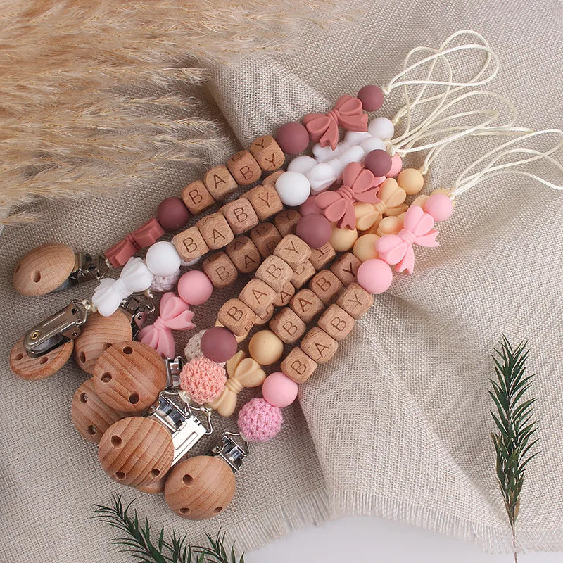 A collection of personalized silicone pacifier clips and a floral-themed lanyard arranged on a beige fabric.