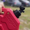 A hand is holding a red plastic faucet adapter for a fuel can, showing the black tap mechanism.