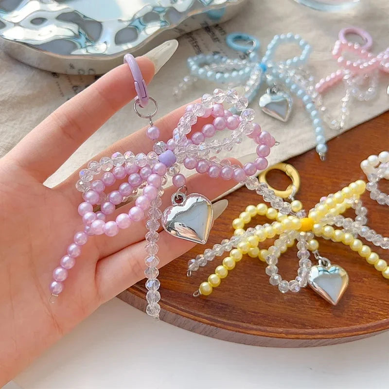 A hand holds a pink and purple pearl bow keychain with heart charms, displayed on a wooden surface.