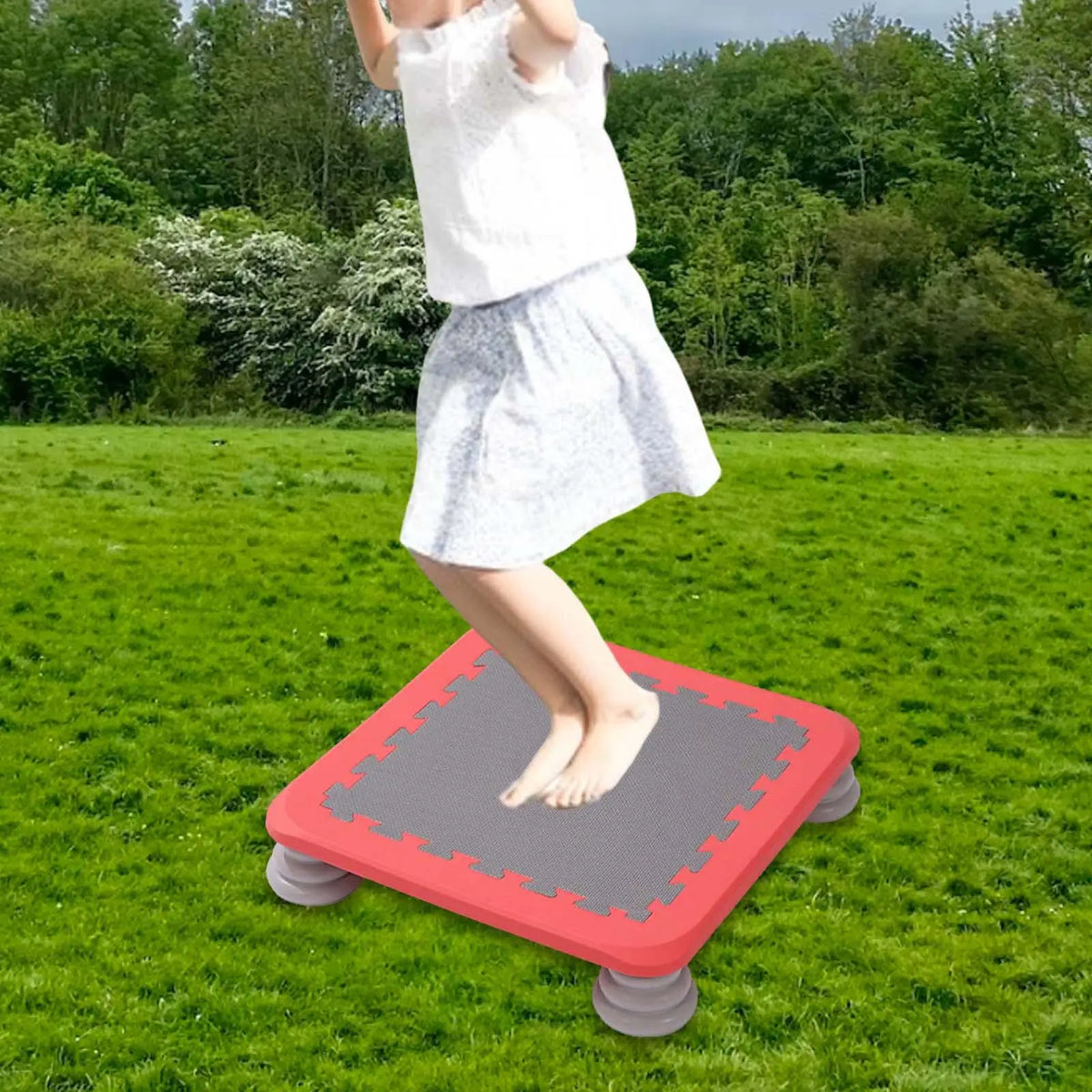 A child is standing on a red mini trampoline outdoors in a grassy field.