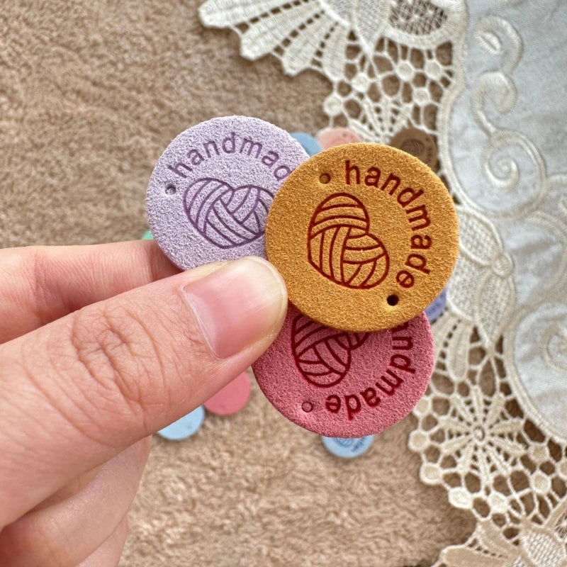 A hand is holding several round, handmade PU leather labels in shades of purple and orange on a beige fabric background.