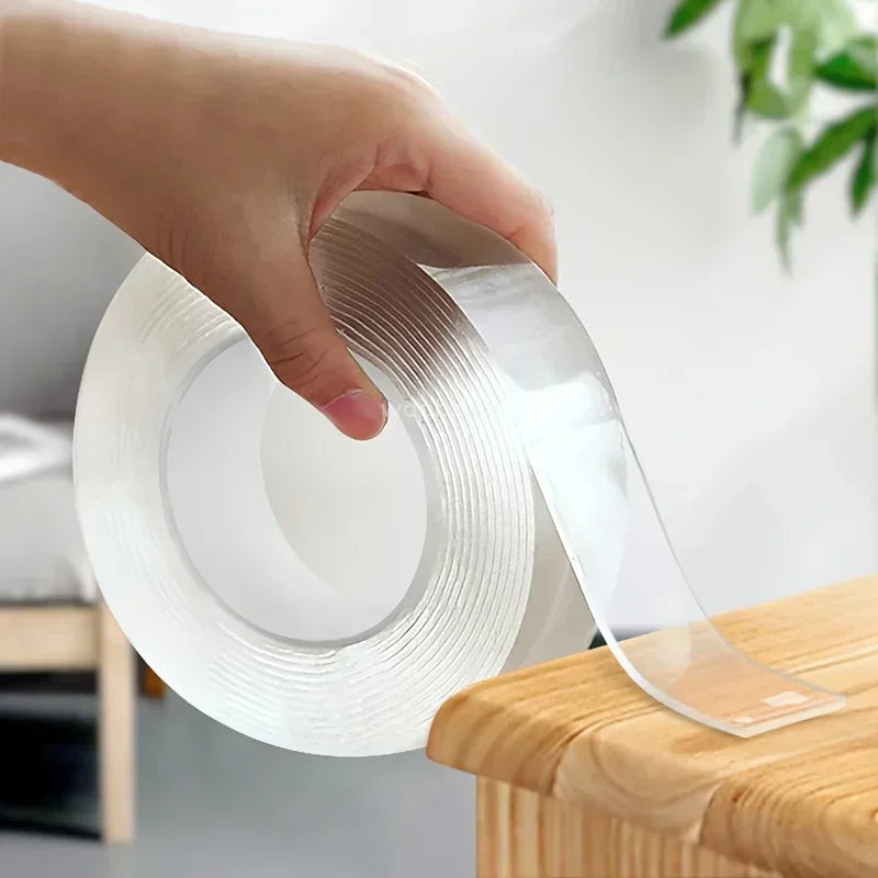 A hand is holding a roll of clear, double-sided adhesive tape on a wooden surface.