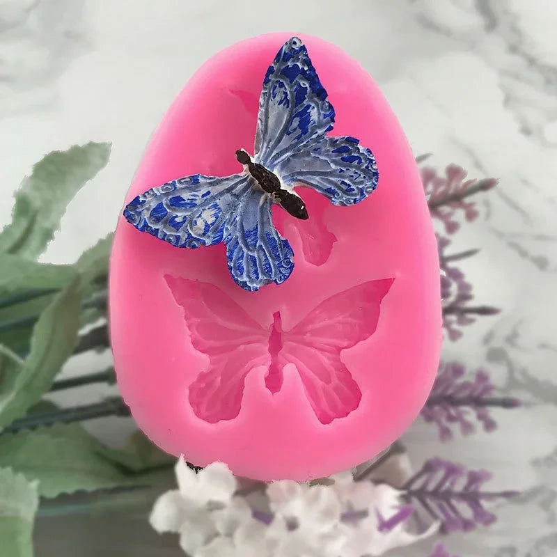 A pink silicone mold shaped like a butterfly is displayed on a white surface with green foliage.