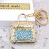 A gold-toned handbag keychain decorated with blue crystal rhinestones, set against a white background.