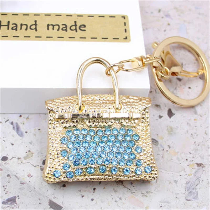 A gold-toned handbag keychain decorated with blue crystal rhinestones, set against a white background.