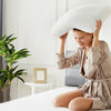 A woman is holding a white memory foam pillow with cooling ventilation and a removable cover in a bright bedroom setting.