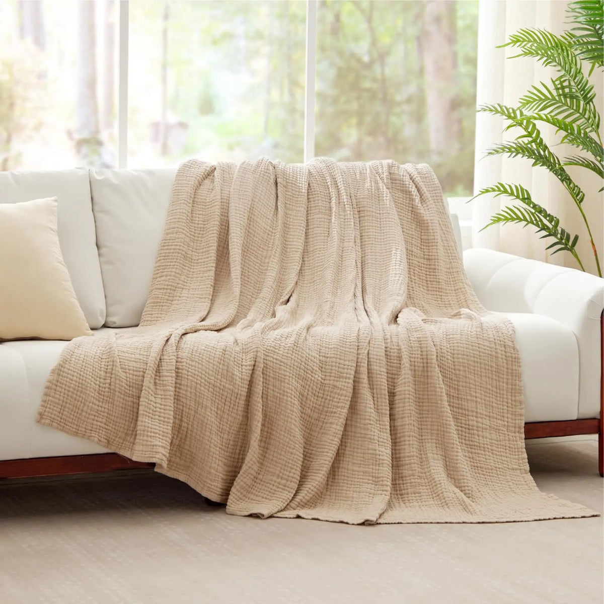 A beige soft muslin cotton blanket draped over a white sofa near a window.
