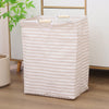 A beige striped foldable laundry basket with light wood handles sits on a wooden floor.
