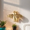 Golden bee-shaped resin wall decor mounted on a white wall next to potted plants.