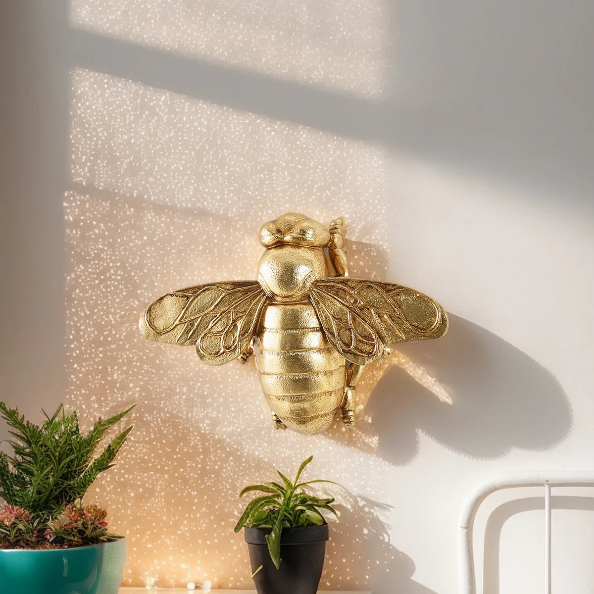 Golden bee-shaped resin wall decor mounted on a white wall next to potted plants.