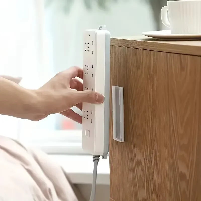 A hand is attaching an adhesive cable organizer to a wooden cabinet, showing the white device and its cable.