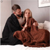 A woman is relaxing on a bed with a man, wrapped in a cozy brown waterproof throw blanket.