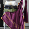 A deep purple luxury bath towel set with gold accents is draped over a white stand, stacked with folded towels.