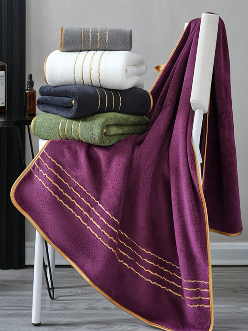 A deep purple luxury bath towel set with gold accents is draped over a white stand, stacked with folded towels.
