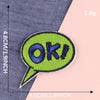 A bright lime green embroidered speech bubble patch with the text OK! on a dark blue fabric background.