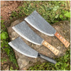 A forged steel axe with a wooden handle rests on a rock amidst green foliage.