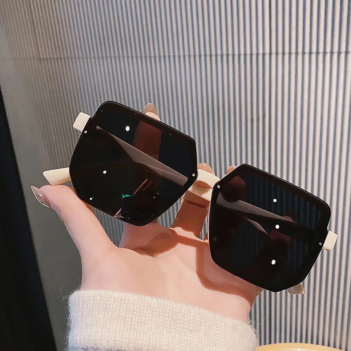 Una mano sostiene unas gafas de sol cuadradas negras vintage contra un fondo a rayas.