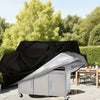 A silver waterproof barbecue grill cover is shown open on a patio with green foliage in the background.