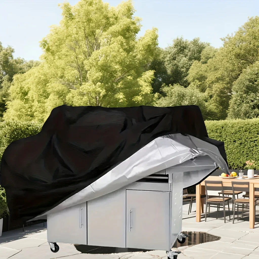 A silver waterproof barbecue grill cover is shown open on a patio with green foliage in the background.