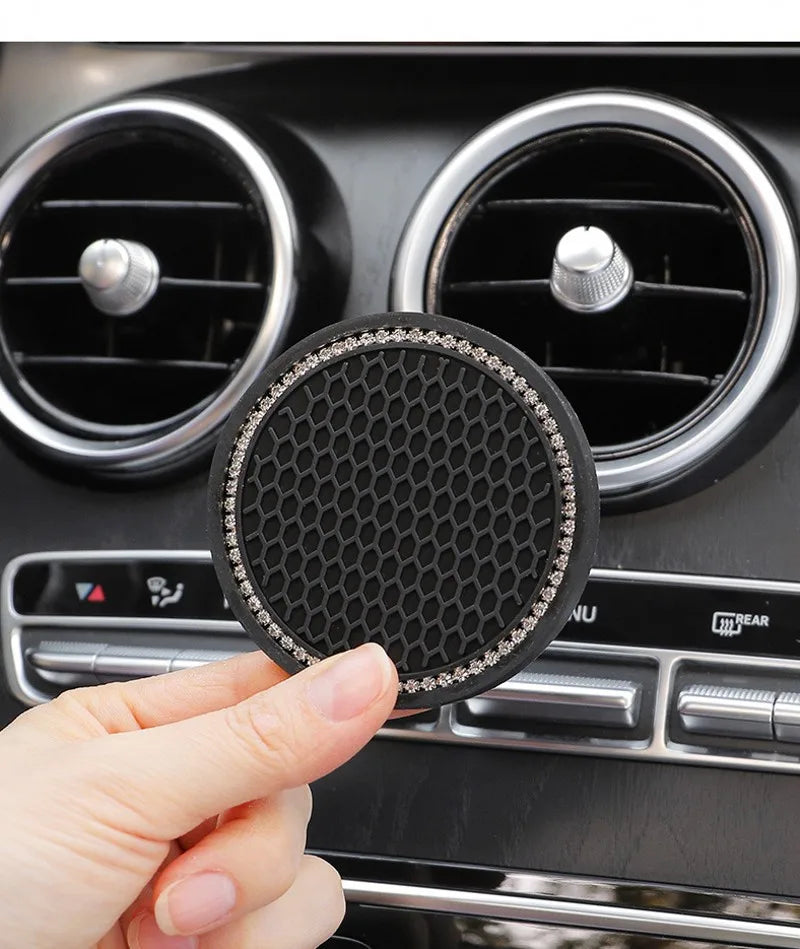 A hand holds a black crystal rhinestone non-slip cup mat for cars, positioned in front of a car stereo.
