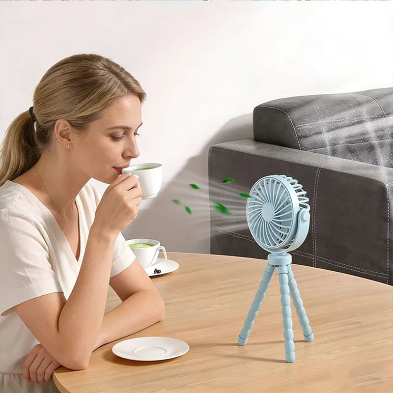 A woman is holding a white cup while sitting at a wooden table next to a blue flexible octopus USB fan on a tripod stand.