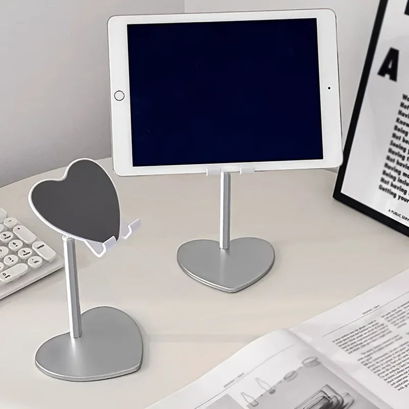 A white adjustable heart-shaped stand holding a tablet and a phone on a light surface.