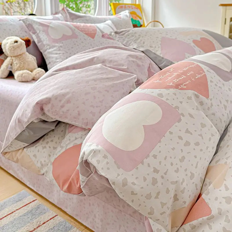 A set of pink and gray cotton duvet covers and pillow shams with heart patterns and a teddy bear decoration.