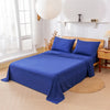A deep blue bedding set featuring a fitted sheet, flat sheet, and two pillows displayed on a wooden bed in a bright room.