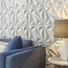 A gray sofa is positioned in front of a textured, geometric white wall panel design with a lamp on a side table.