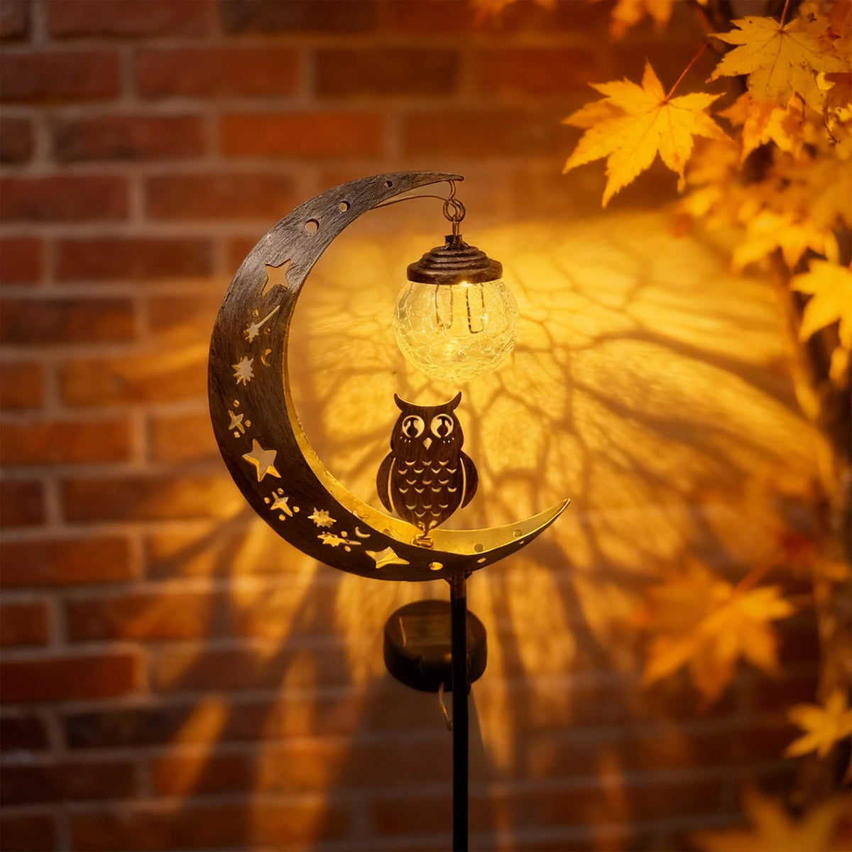 A solar moon-shaped garden light featuring an owl silhouette and a glass ball, casting shadows on a brick wall.