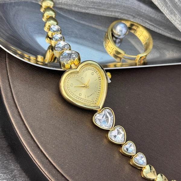 Goldfarbener herzförmiger Quarz-Armband mit klaren Steinen und eine goldene Uhr auf dunklem Hintergrund.