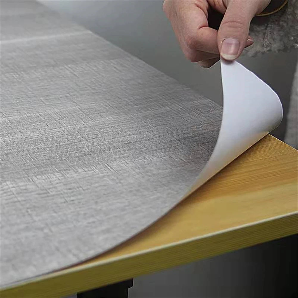 A hand is peeling a sheet of self-adhesive wood grain wallpaper film from a roll onto a wooden surface.