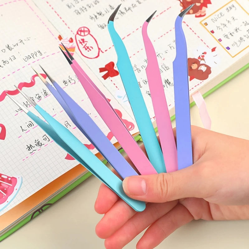 A hand holds several precision craft tweezers with various colored straight and curved tips resting on a notebook page.