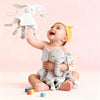 A baby is holding a white elephant plush toy with a teether and comfort blanket against a pink background.