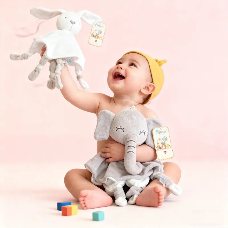 A baby is holding a white elephant plush toy with a teether and comfort blanket against a pink background.
