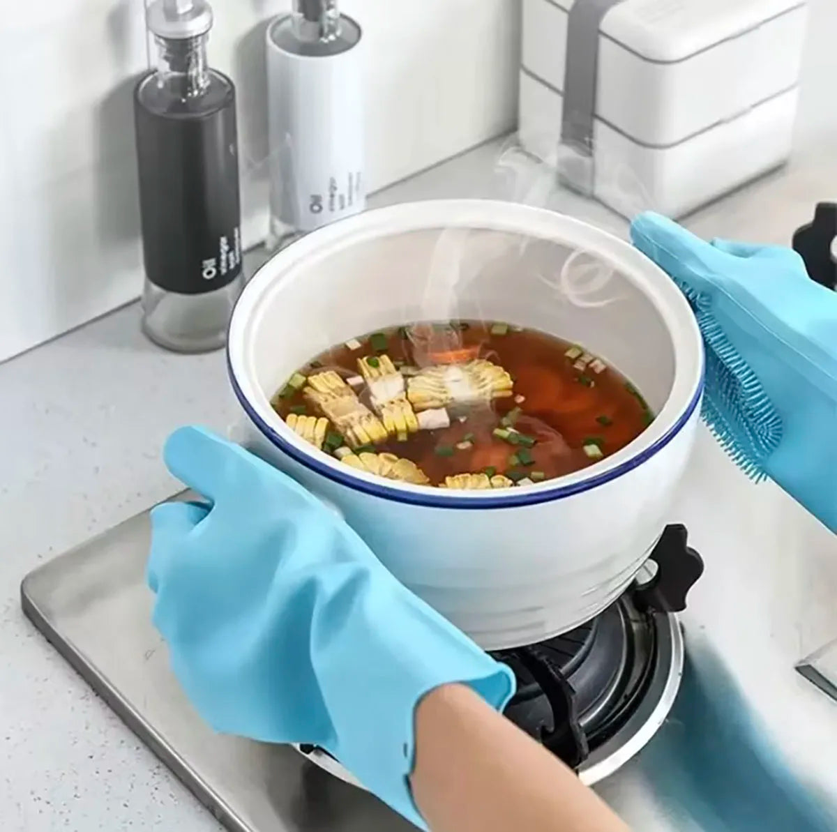 A hand wearing a blue scrubber-textured dishwashing glove is holding a white bowl of soup on a stovetop.
