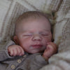 A newborn baby doll with closed eyes resting in a light-colored fabric.
