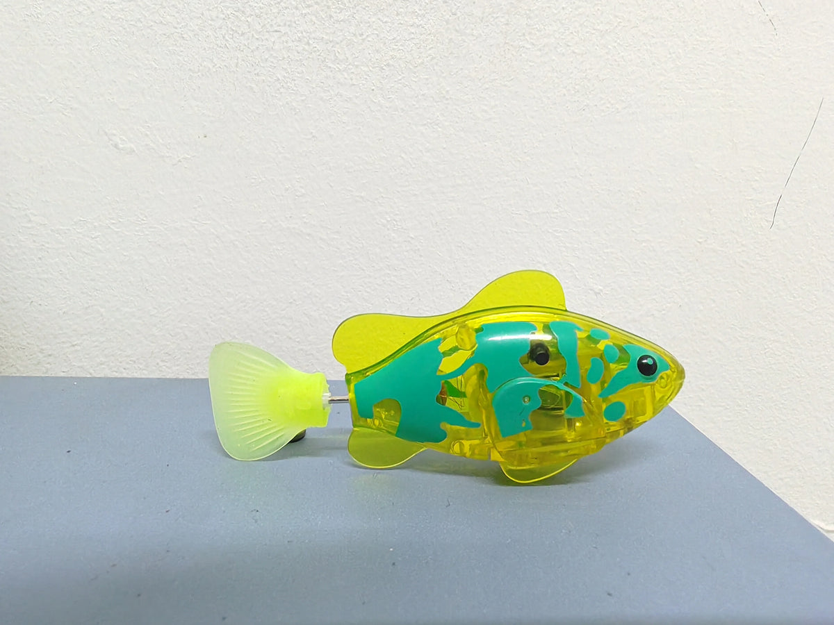 A yellow and teal light-up sensor swimming fish toy resting on a gray surface.