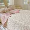 A pink floral quilted bedspread with white accents, featuring a pink knitted throw and stuffed rabbit pillows.